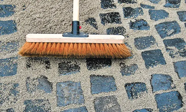 Besen verteilt fugensand gegen unkraut auf einem gepflasterten Weg.